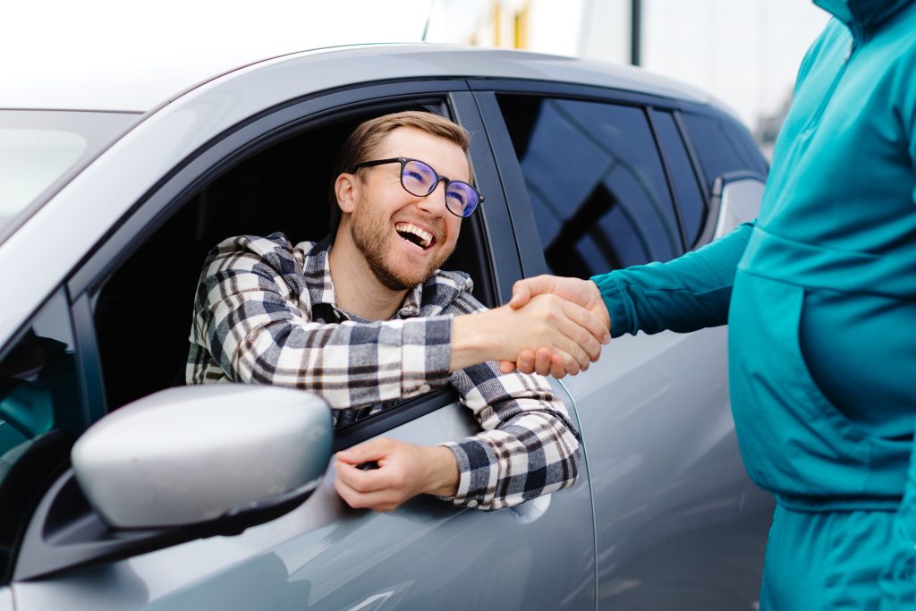A man in a car smiling while shaking hands with someone outside.