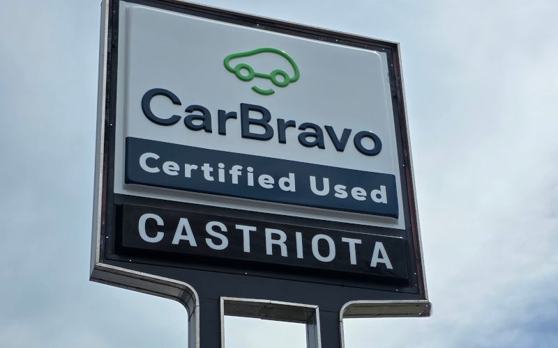 A roadside CarBravo Certified Used sign by Castriota, with a green car icon, against a cloudy sky.